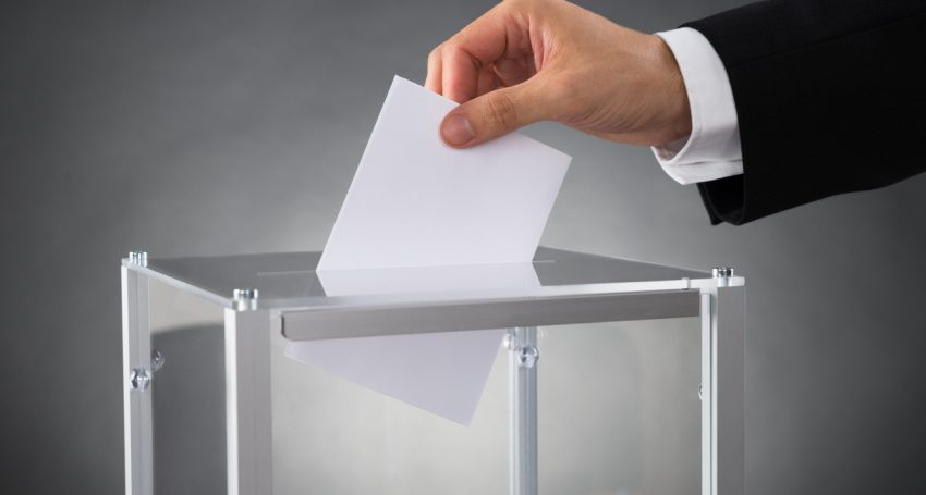 Close-up Of Businessperson Hands Putting Ballot In Box At Desk